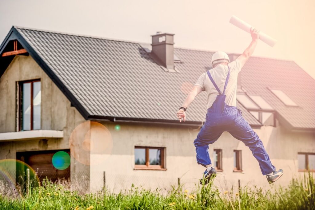 a man in overalls and a white hard hat jumping in the air with a paper roll in his right hand in front of house. Conventional rehab loan, va one time close construction loan